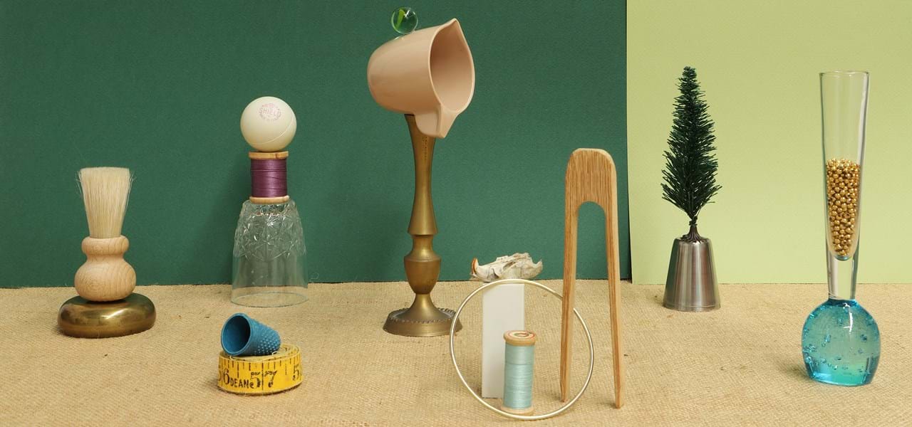 A collection of small objects are arranged against a green background, including a small shaving brush, a toy tree, a blue thimble, reels of cotton, a pair of wooden tongs and a yellow measuring tape. Some of the objects are balancing on top of one another. 