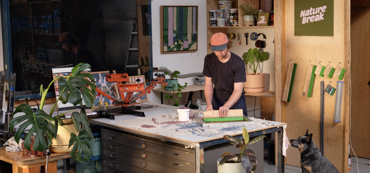 A man wearing a baseball cap bends over a bench at which he is screen printing. He is in a studio and surrounded by printmaking equipment, plants and artworks. A sign that says Nature Breaks hangs on the wall behind him and a blue heeler sits beside the bench and stares up at him adoringly.