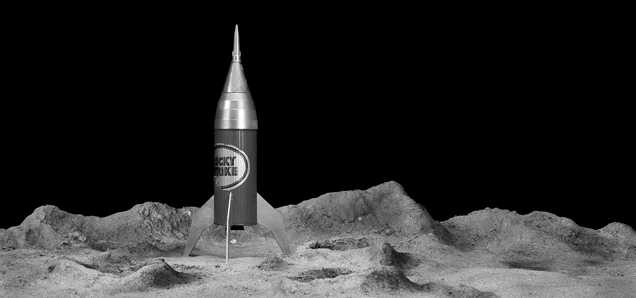 A black and white photograph of a space rocket sitting on a sandy surface that looks like the moon.